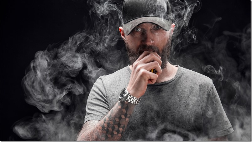 bearded male dressed in a grey shirt, sunglasses and baseball cap vaping