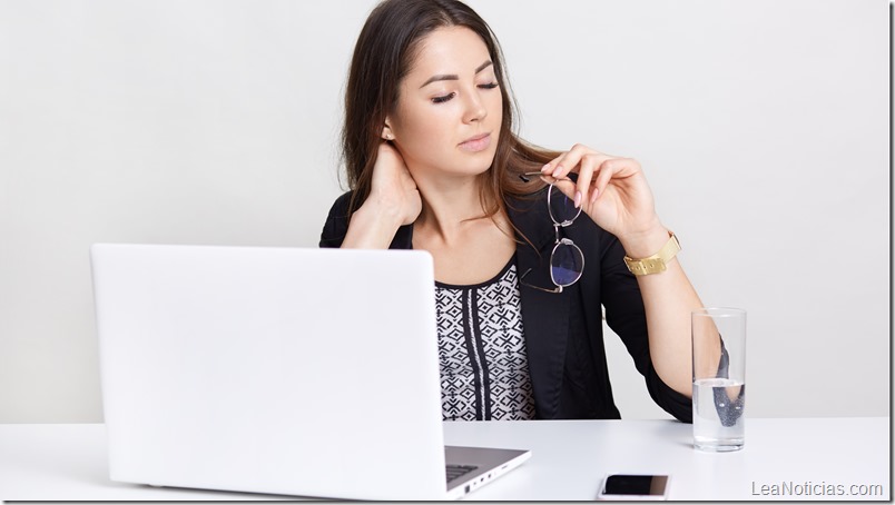 Image of tired Caucasian woman works long hours on portable laptop computer, holds spectacles in hand, feels pain in neck, surrounded with modern technologies, sits at white desk, poses indoor