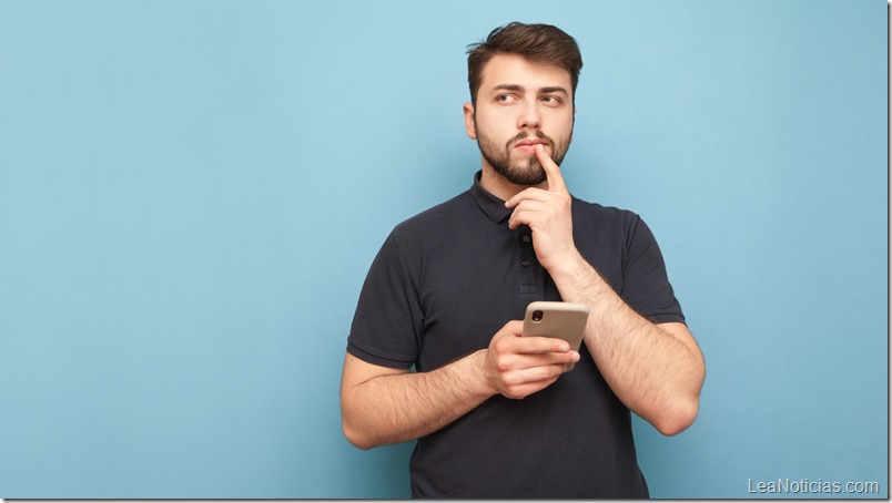 Thoughtful man with a beard standing on a blue background with a smartphone in his hand, looking sideways and thinking wearing a dark t-shirt. Isolated. Copy space